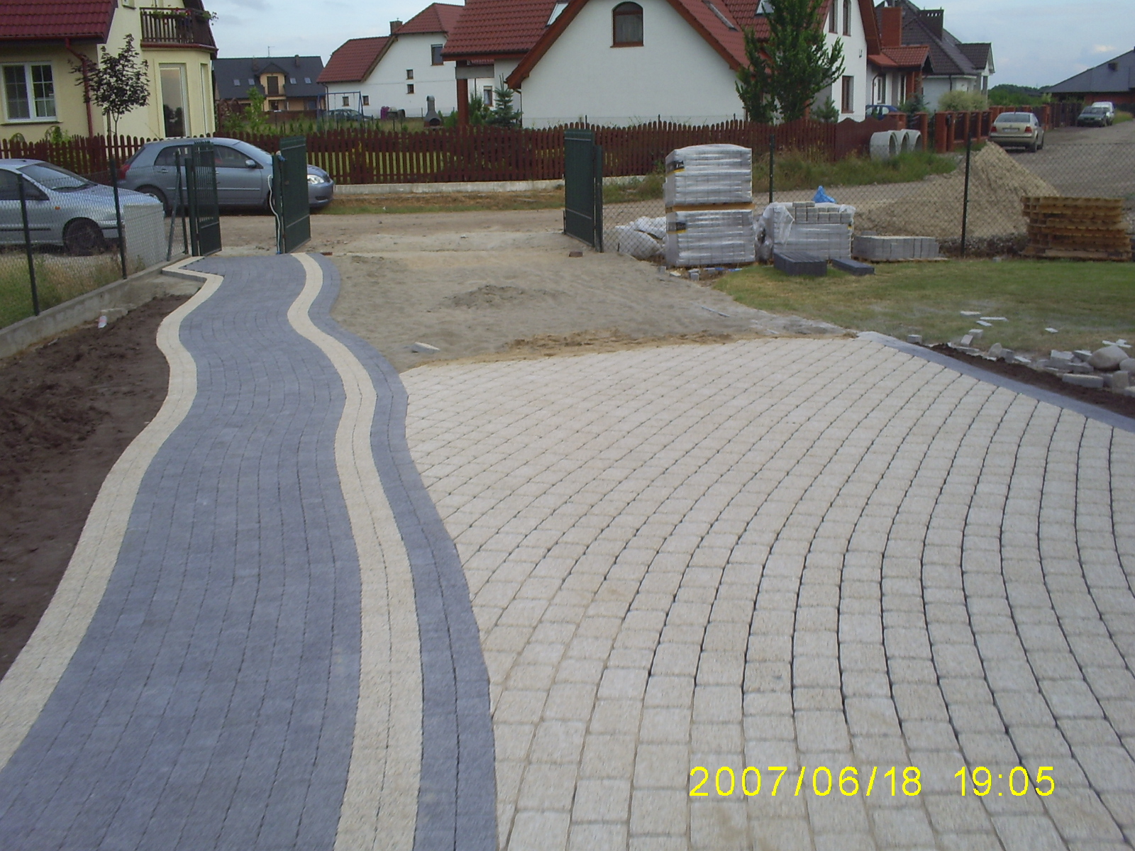 Ułożona nawierzchnia z kostki brukowej w dwóch odcieniach szarości, tworząca falujący wzór ścieżki i prostej powierzchni, z widocznymi zapasami kostki w tle i domami jednorodzinnymi.